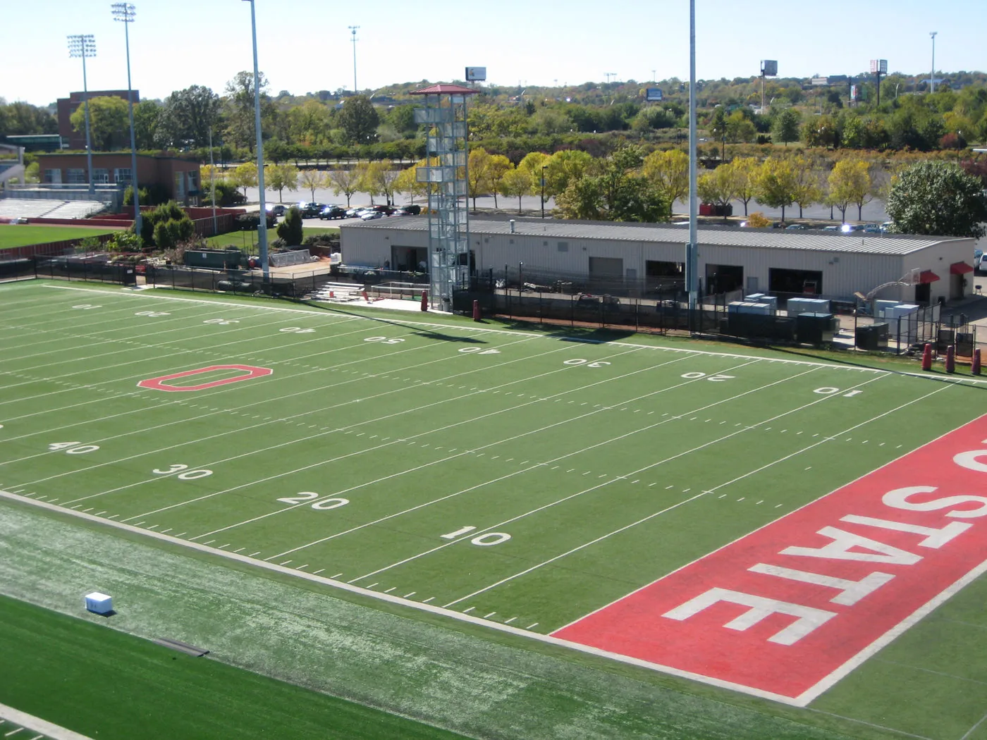 Aerial view of an Ohio State University practice football field with a large red end zone reading OHIO STATE, an athletic support building, and a parking area in the background.