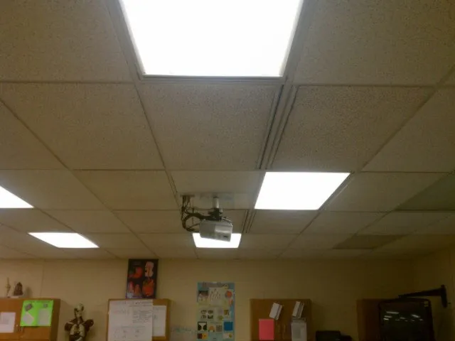 Classroom ceiling with a suspended-tile grid, recessed lighting, and a ceiling-mounted projector positioned above a teacher's desk area. (Briggs School)
