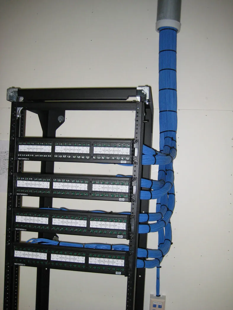 Two-post equipment rack loaded with four NextSpeed Cat6 patch panels and a bundle of blue cabling entering through an overhead conduit.