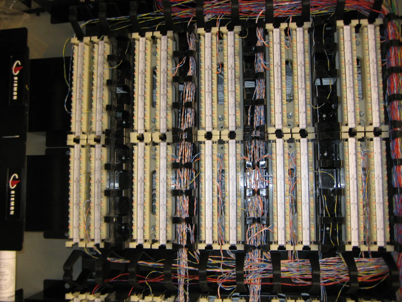 Wall-mounted telecommunications rack with rows of Siemon patch panels and bundled white and green cable trunks feeding from an overhead cable tray.