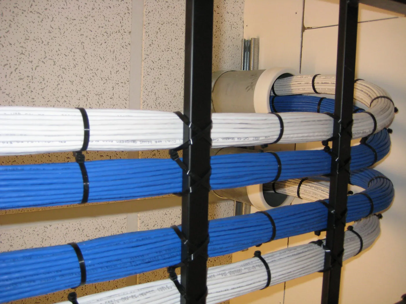 Bundled blue and white network cabling secured with cable ties and routed through a two-post equipment rack toward a wall conduit.