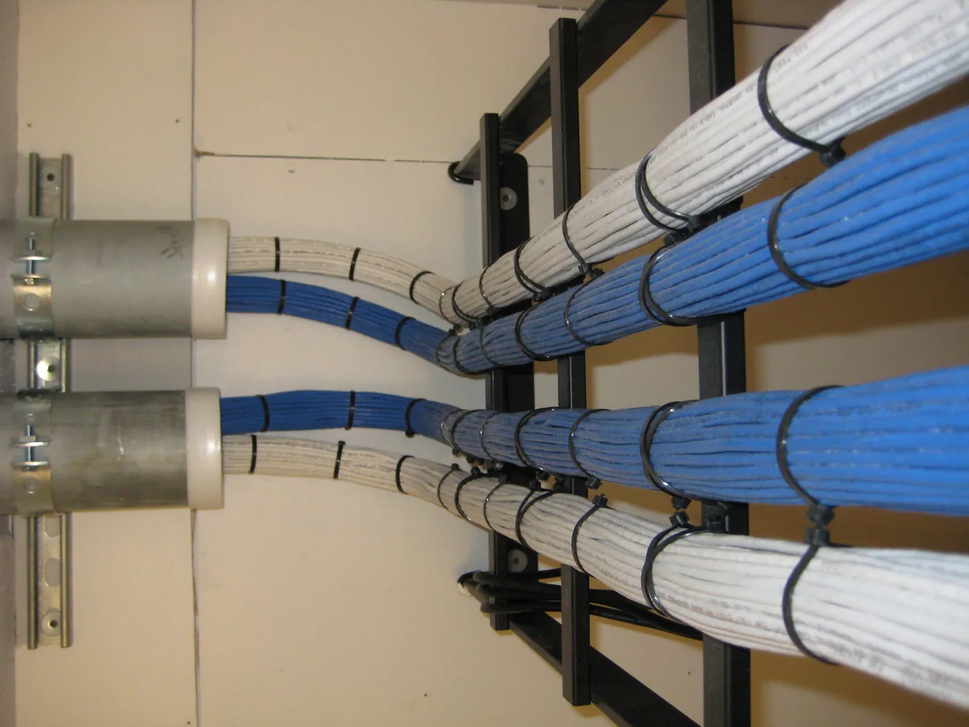 Bundled blue and white network cabling routed through a two-post rack and around vertical cable rings, feeding into wall conduit above.