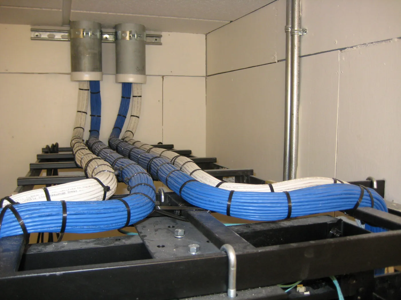Bundled cable runs feeding down from two ceiling conduits into a two-post equipment rack in a school telecom closet.