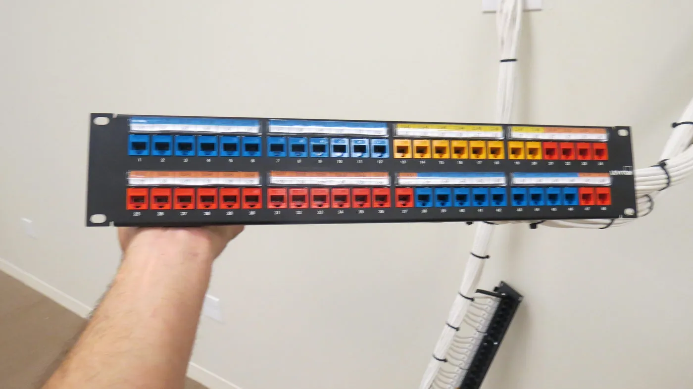 Technician holding an open patch panel loaded with blue, orange, red, and yellow RJ45 network jacks.