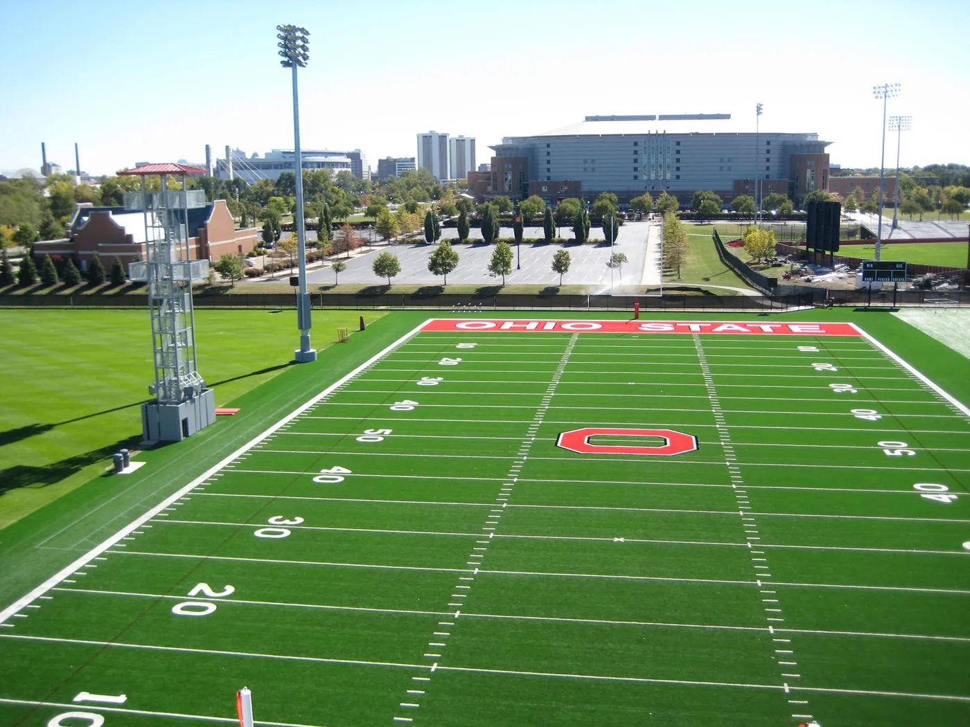 View across an Ohio State University practice football field toward a large campus arena building, a parking lot, and a tree-lined walkway.