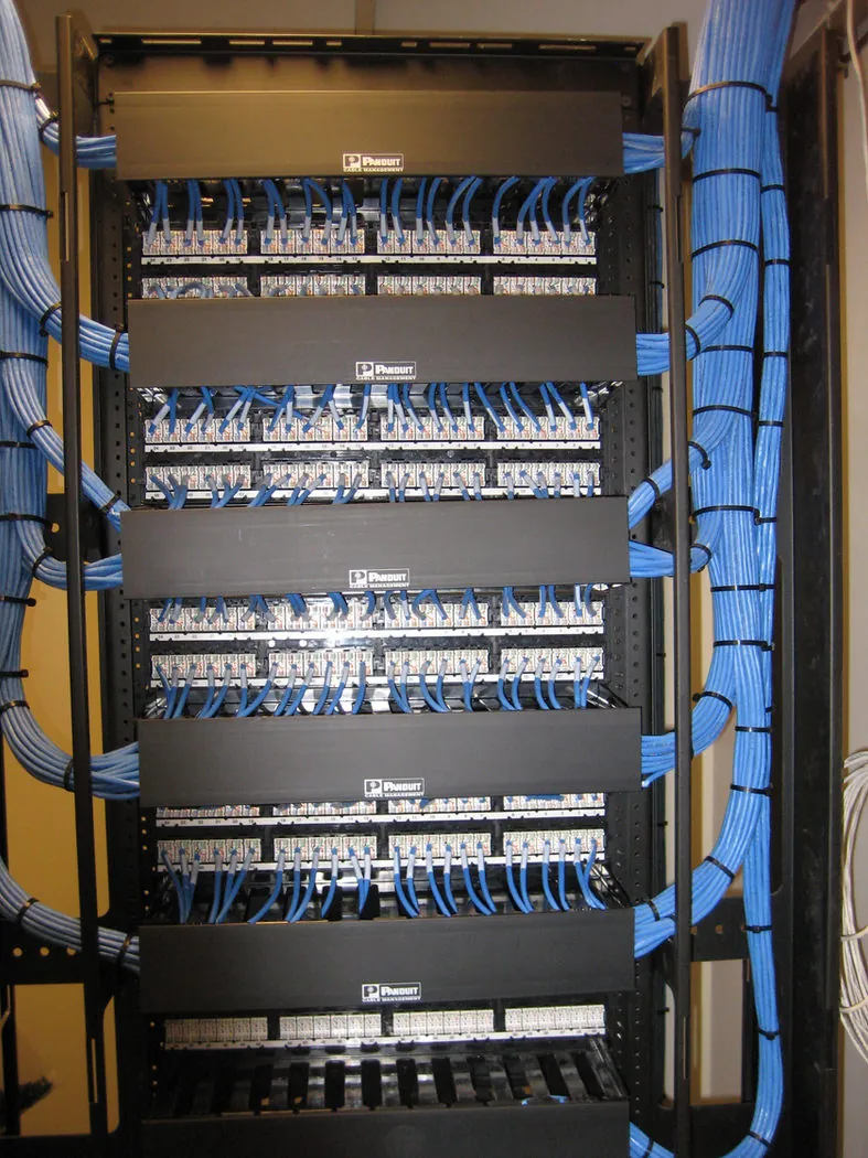 Bundled blue and white network cabling routed along a ceiling cable rack, feeding down through conduit sleeves into an equipment rack.