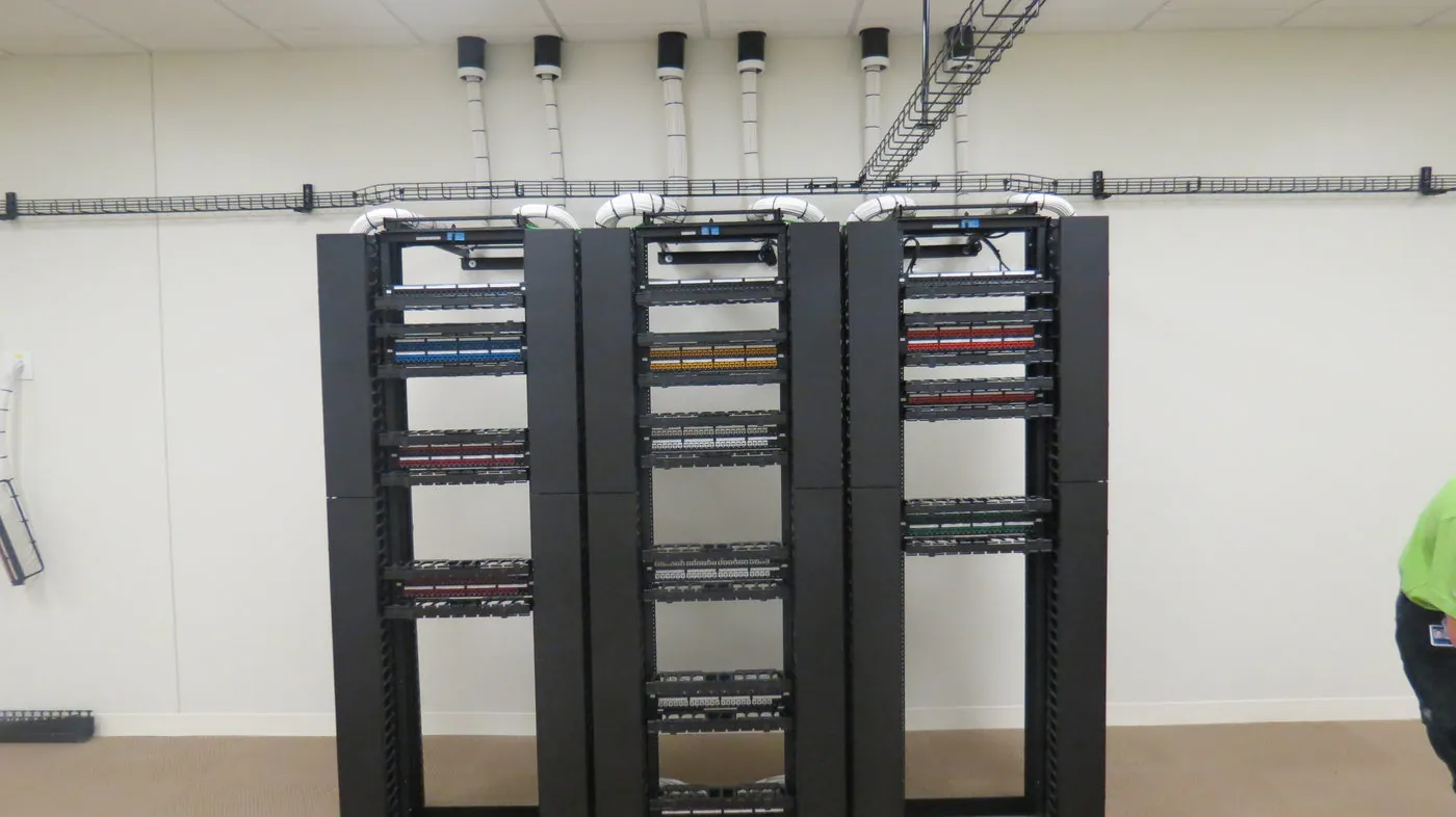 Three open equipment racks loaded with color-coded patch panels, fed by an overhead cable tray running along the ceiling.